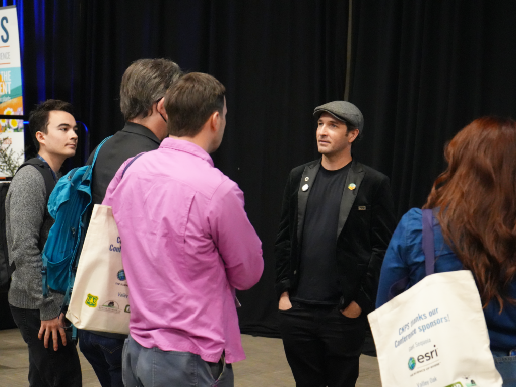 Man in a black blazer and gray flat cap talks to a small group at an event, with dark stage curtains in the background.