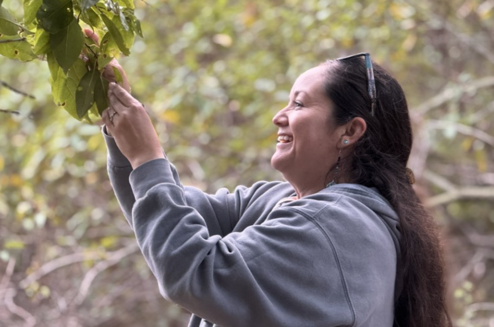 Woman smiling while reaching for leaves in a green, outdoor setting, wearing a gray hoodie and sunglasses on her head.