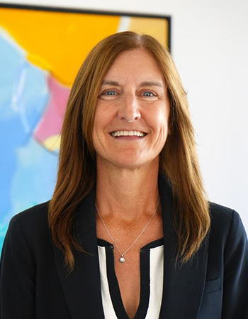 Professional portrait of a smiling woman with shoulder-length brown hair, wearing a black blazer and white blouse, standing in front of a colorful abstract painting.