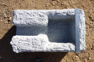 Top-down view of a light-gray concrete block with a hollow cavity, placed on dirt, with a metal ruler laid along its side for scale.