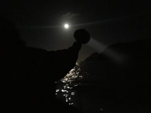 Moonlit night over a dark sea with jagged rocks in the foreground and a luminous moon above, reflecting on the water.
