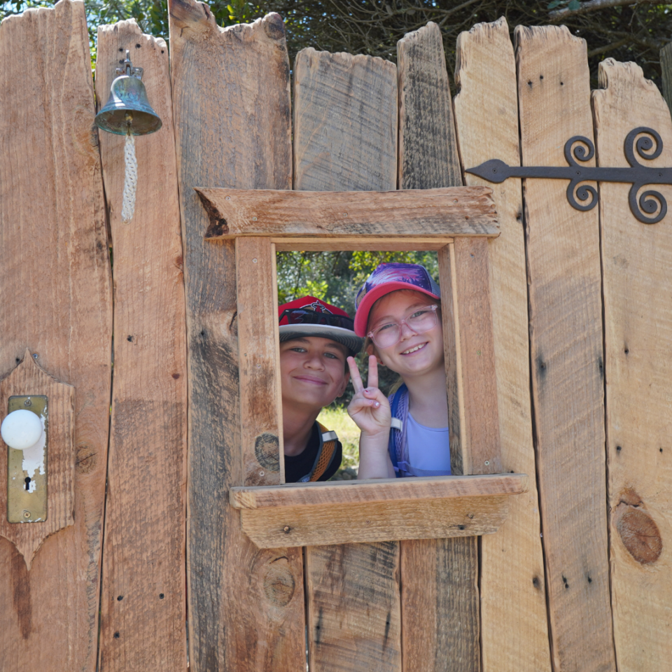 Two kids peek through a rustic wood frame window in a fence, making peace signs and smiling at the camera.