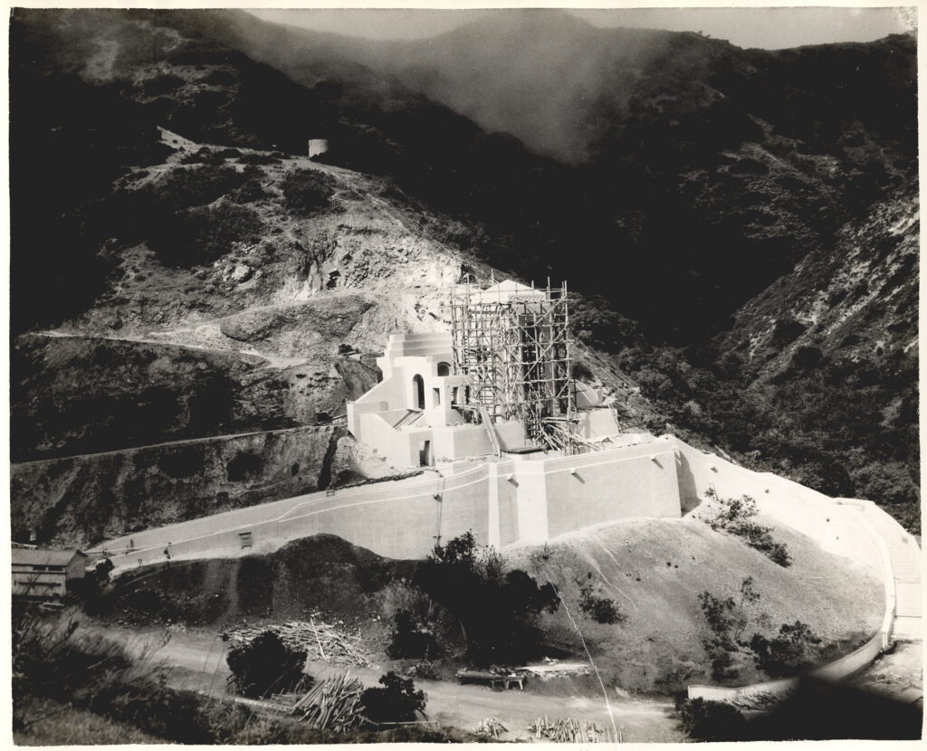 Construction site on a hillside with scaffolding around a white angular building, a winding road nearby, and rugged, shadowed mountains in the distance.