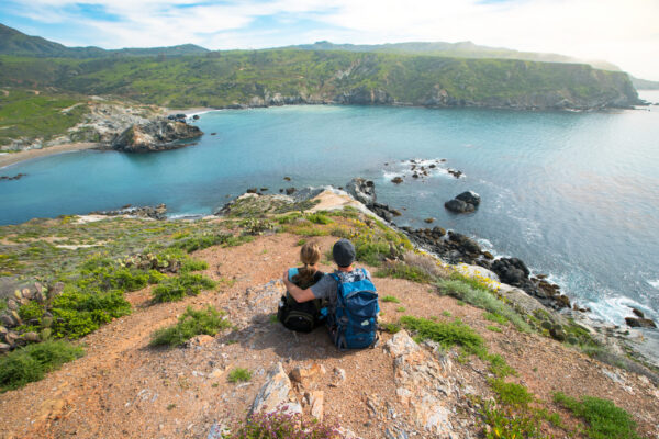Two hikers with backpacks sit on a rocky hillside overlooking a blue cove and green cliffs. A peaceful coastal view.