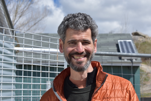 Portrait of a smiling man with gray hair and beard, wearing an orange jacket outdoors.