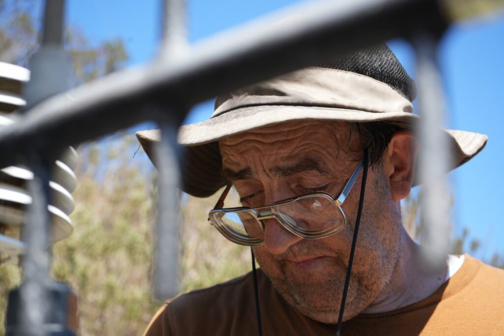 Older man wearing a beige bucket hat and glasses looks downward outdoors, with blurred fence bars in the foreground.