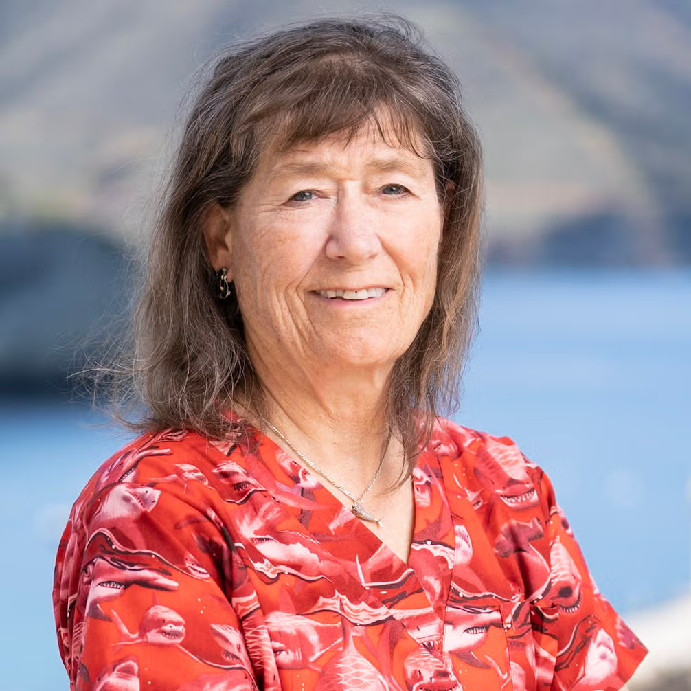 Portrait of an older woman with shoulder-length brown hair, smiling, wearing a red patterned blouse outdoors by a blue body of water.