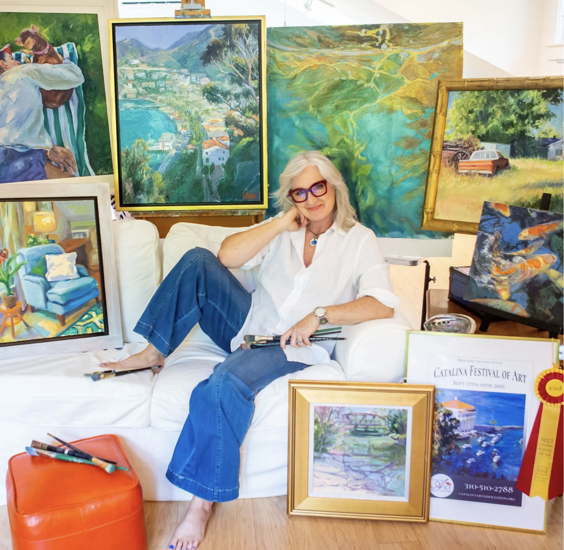 Woman with white hair and red glasses sits on a white couch among vibrant landscape paintings and framed artwork in an art studio/sale space.