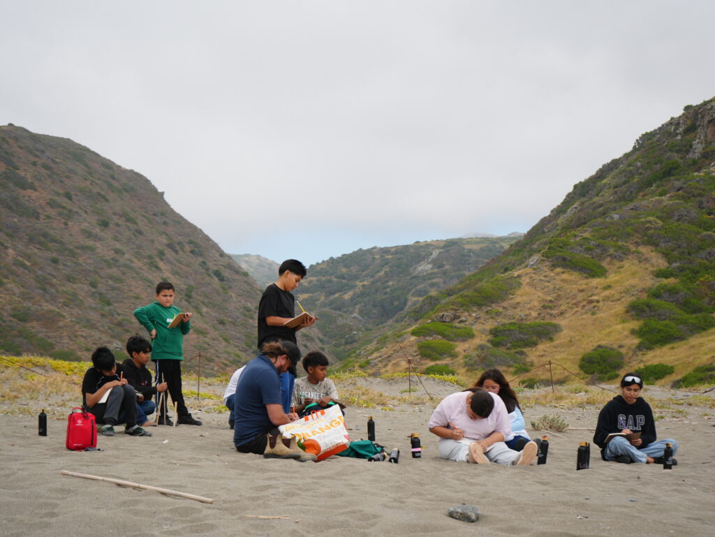 Group of students sitting on a sandy beach, writing in notebooks with bags and water bottles nearby, mountains in the background under a cloudy sky.