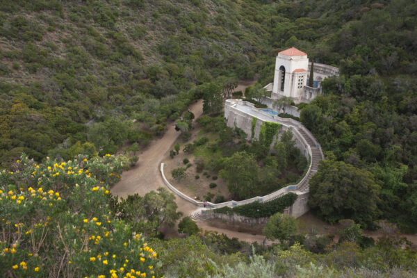 A white, tower-like building with a red-tiled roof perched on a terraced hillside surrounded by dense trees and a curved pathway below.