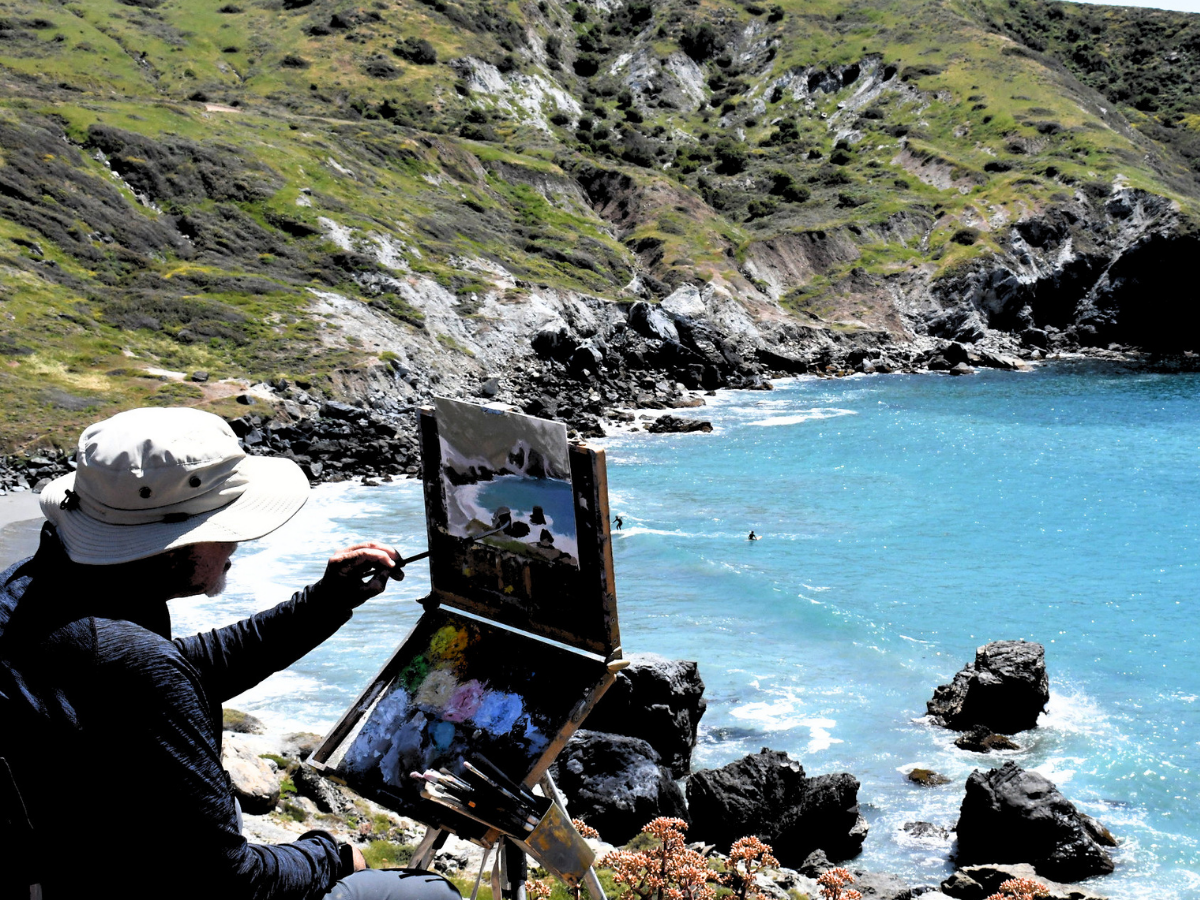 Artist wearing a white hat paints at an outdoor easel by a rocky, turquoise bay surface.