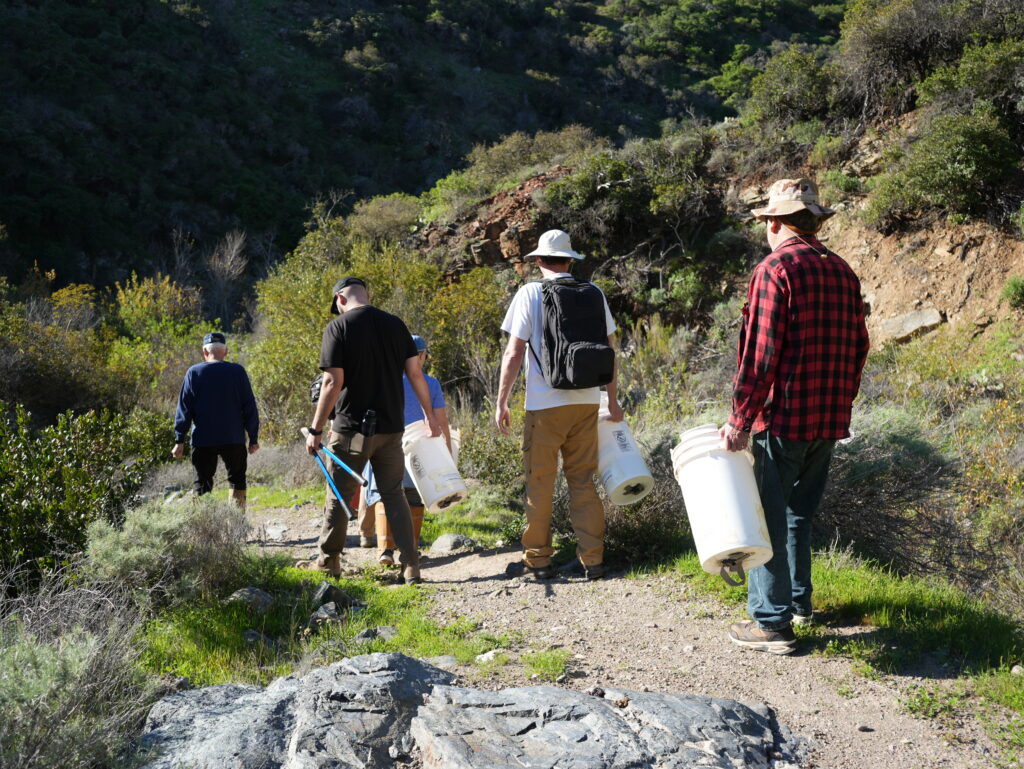 Group of hikers walking along a rocky trail, carrying large white buckets, in a hillside canyon setting.