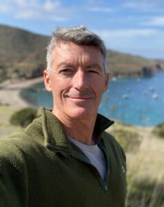 Close-up portrait of a man in a green fleece jacket, smiling, with a blue lake and hills in the background on a sunny day.